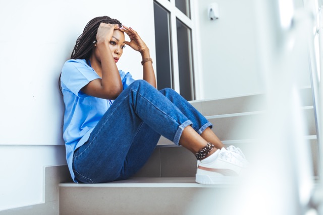 Tired exhausted female african scrub nurse wears face mask blue uniform gloves sits on hospital floor. Depressed sad black ethic doctor feels fatigue burnout stress, lack of sleep, napping at work